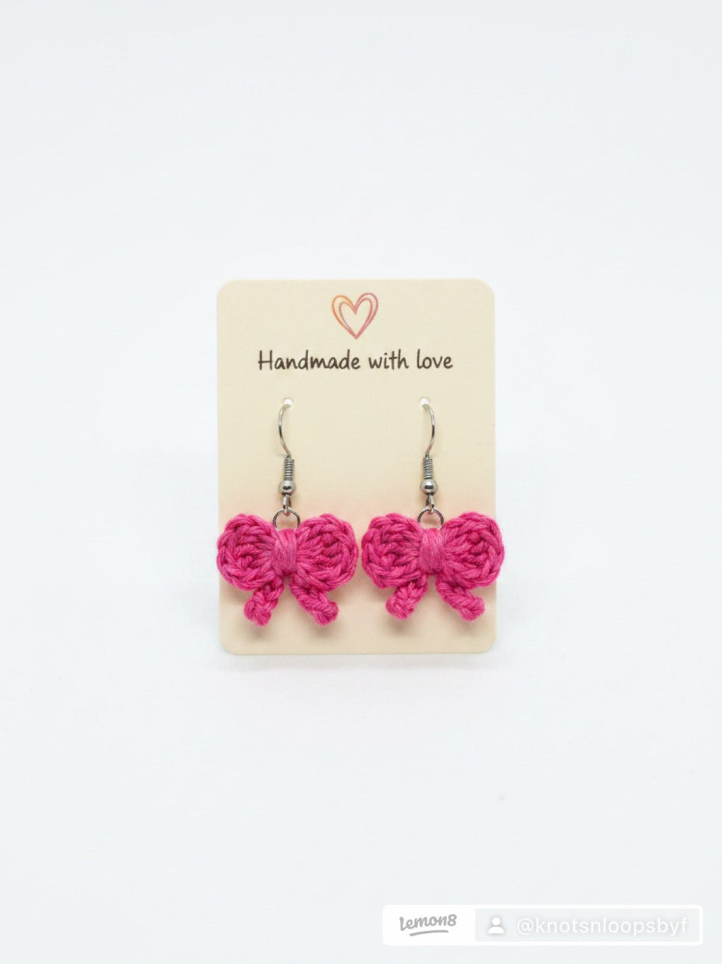 A pair of bright pink crocheted bow dangle earrings on a beige display card that reads 'Handmade with love' against a plain white background.