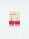 A pair of bright pink crocheted bow dangle earrings on a beige display card that reads 'Handmade with love' against a plain white background.