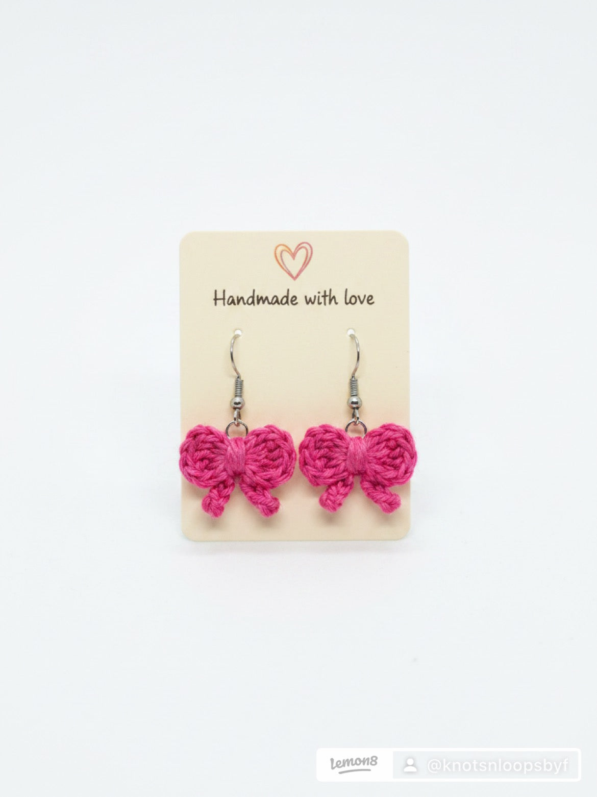 A pair of bright pink crocheted bow dangle earrings on a beige display card that reads 'Handmade with love' against a plain white background.