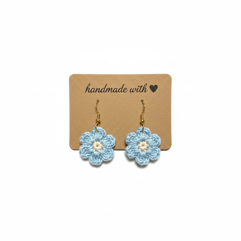 A pair of handmade crochet flower earrings in light blue and white, hanging from gold hooks on a brown kraft display card that reads 'handmade with'.