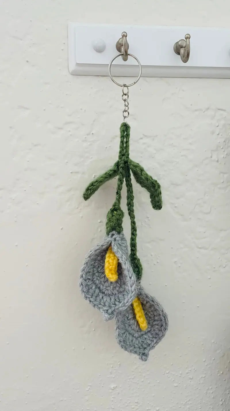 A handmade crocheted keychain featuring two gray calla lilies with yellow centers and green stems, hanging from a metal ring on a white wall hook.