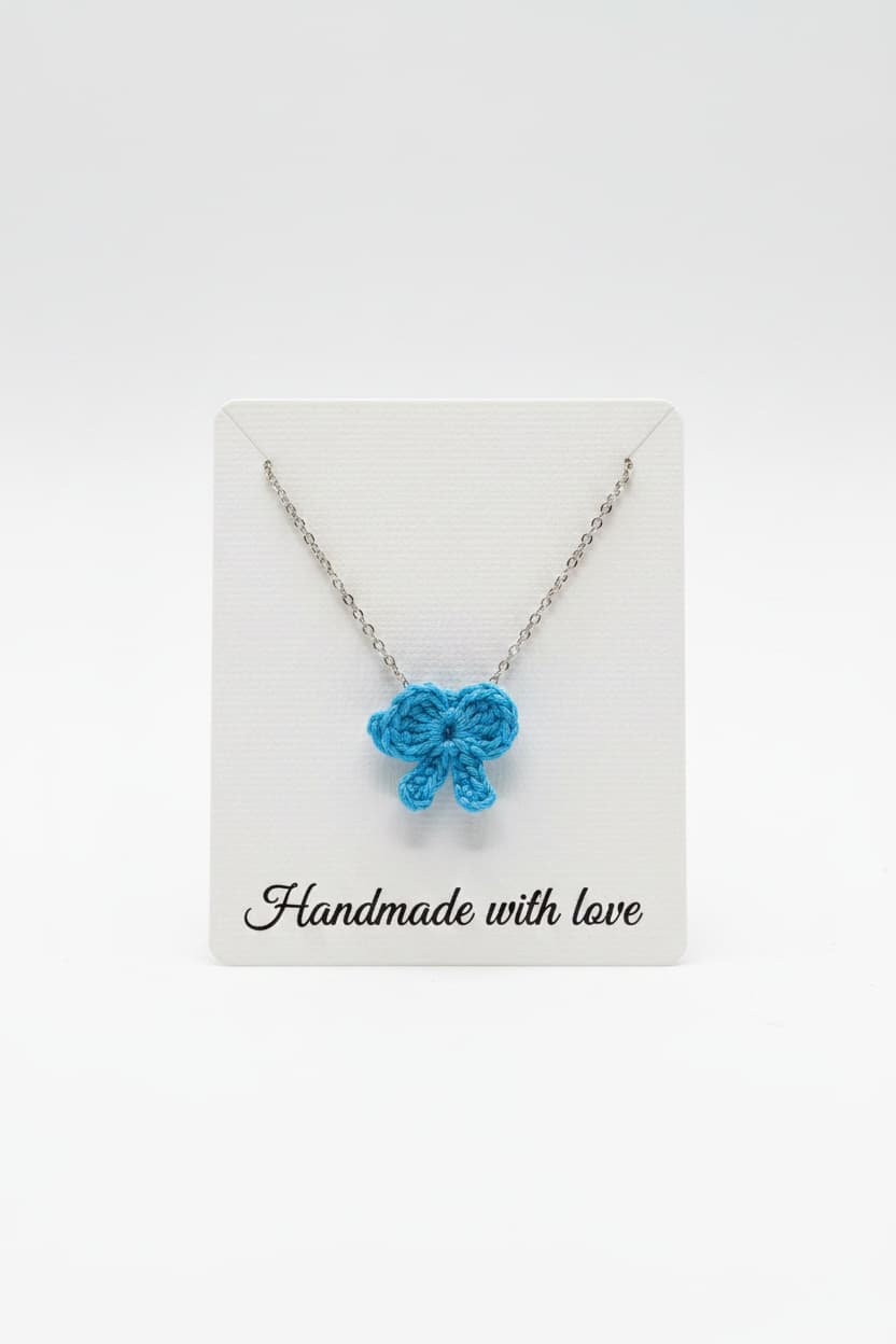 A silver chain necklace with a small crocheted blue bow pendant, displayed on a white card that reads 'Handmade with love' against a white background.