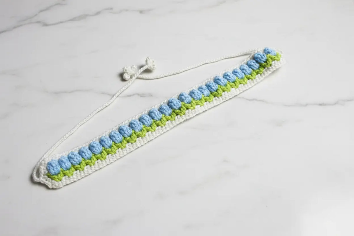 A handmade crochet strap with a white base, featuring rows of light blue and lime green textured stitches, finished with a white adjustable cord and toggle clasp, displayed on a white marble surface.