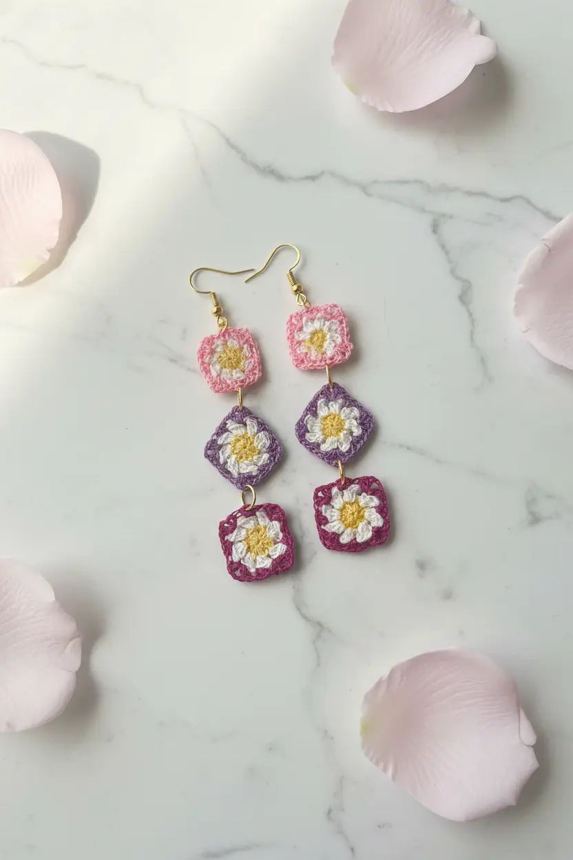 A pair of handmade crochet daisy drop earrings with gold fishhook wires, displayed on a white marble surface with scattered pink rose petals. The earrings feature pink, purple, and white crochet flowers with yellow centers.