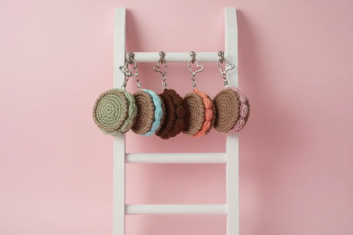 Four crocheted macaron keychains in shades of green, blue, brown, and pink, hanging from a white wooden ladder against a solid pink background.