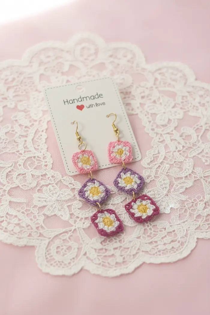 A pair of handmade daisy earrings with gold hooks, pink and purple petals, and white centers, displayed on a white card over a white lace doily on a pink background.