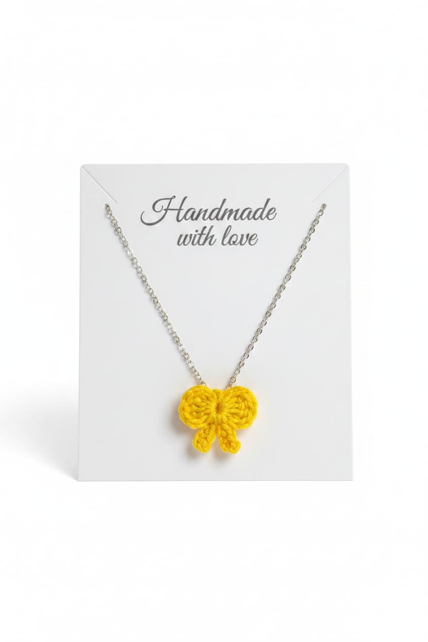 A yellow crocheted bow pendant necklace on a delicate silver chain, presented on a white display card with the text 'Handmade with love' against a clean white background.