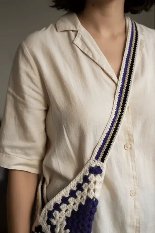 A close-up of a person wearing a cream linen shirt and carrying a crocheted shoulder bag with a cream, purple, and black patterned strap.