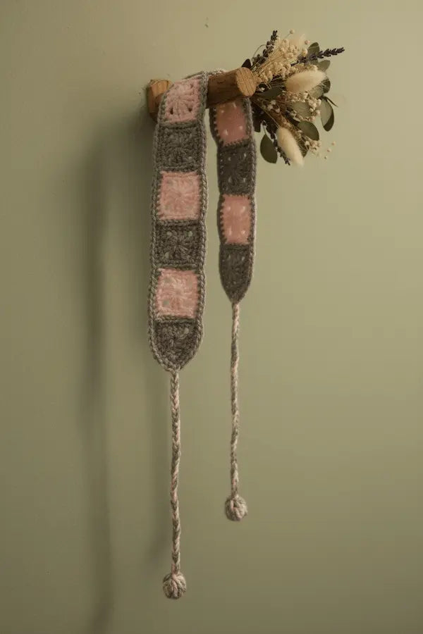 A handmade knitted belt with pink and gray square patches hanging from a wooden peg on a sage green wall, next to a dried floral arrangement.