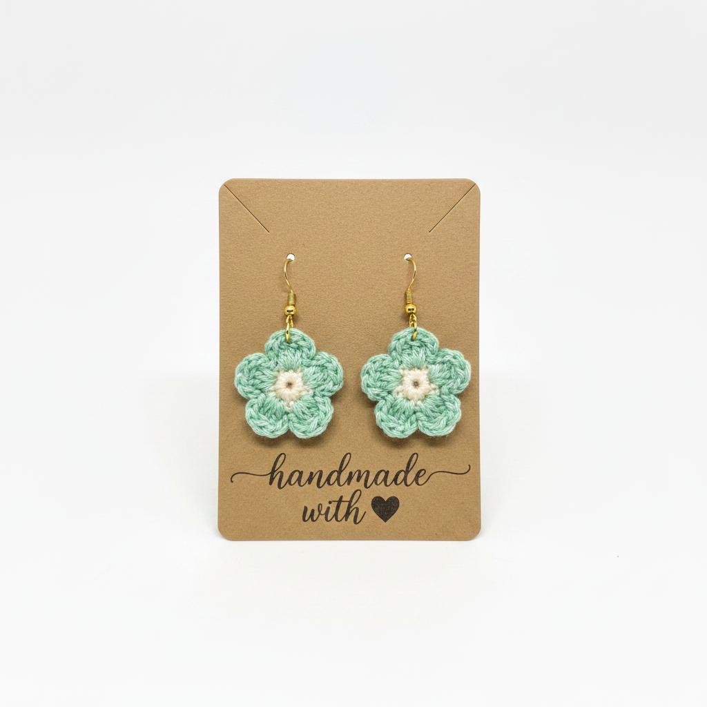 A pair of handmade crocheted flower earrings in mint green with white centers and gold hooks, displayed on a brown kraft card that reads 'handmade with a heart' against a white background.