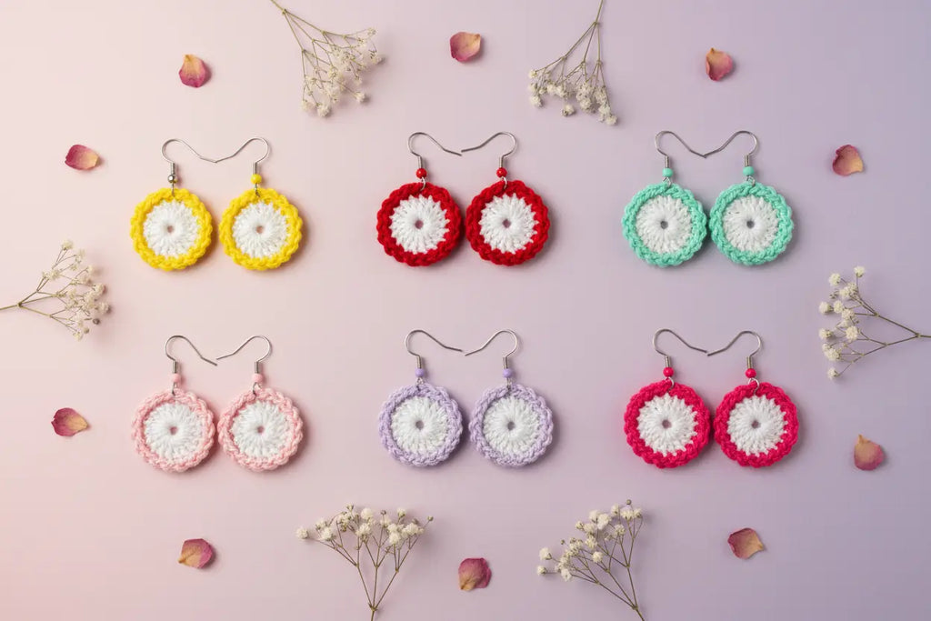 Eight pairs of handmade crocheted dangle earrings arranged in two rows on a pastel pink background with scattered baby's breath flowers and rose petals. The earrings feature circular crochet tops in yellow, red, mint green, pink, lavender, and magenta, with white centers and silver fishhook ear wires.