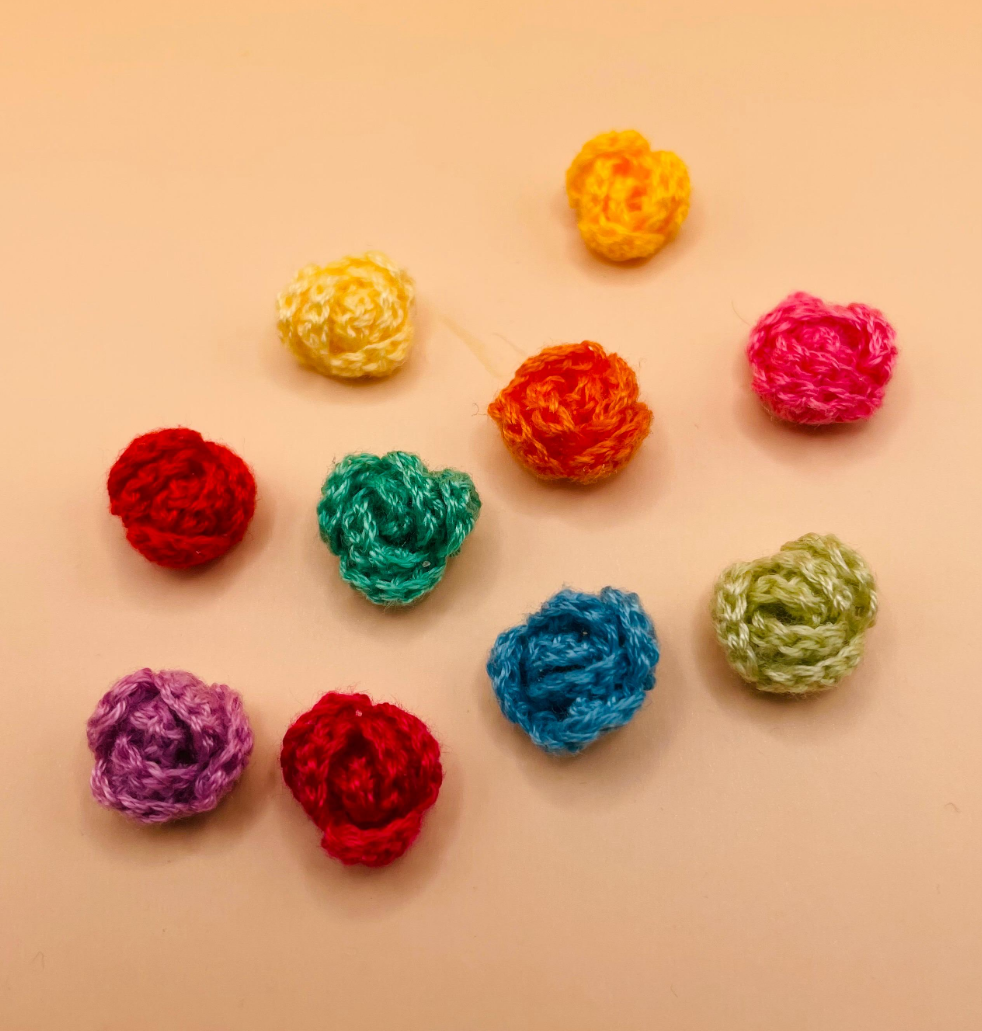 A screenshot of a mobile app showing a photo of ten crocheted rose flowers in various spring colors on a peach-colored background. The screenshot includes interface elements like 'Edit', 'Photo filters', 'Alt text', and a description of the colors of the flowers.