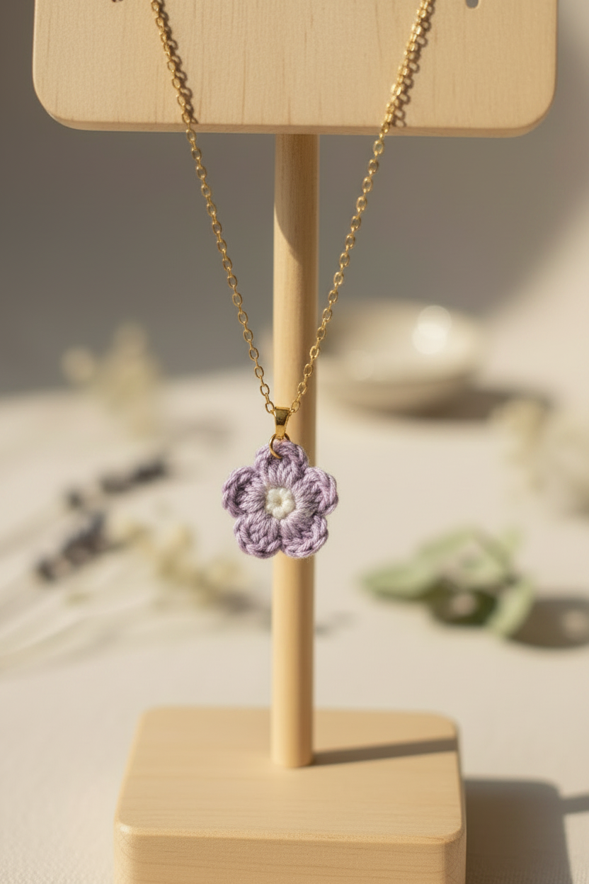 A gold chain necklace with a small crocheted lavender and white flower pendant, displayed on a light wood stand against a softly blurred warm background.