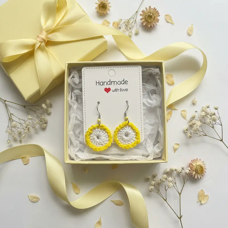 A pair of handmade crocheted yellow and white dangle earrings displayed on a white card inside a square gift box with tissue paper, surrounded by a pale yellow ribbon, a matching gift box, and dried flowers on a white background.