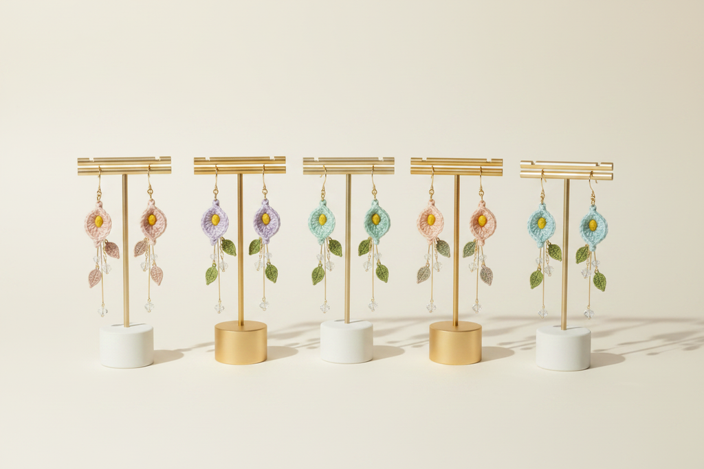 Six pairs of dangling floral earrings displayed on gold T-stands with white and gold cylindrical bases, arranged in a row against a warm off-white background.