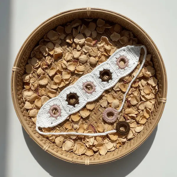 A white crocheted bracelet with pink and brown flower motifs, displayed on a bed of dried flower petals inside a round woven basket.