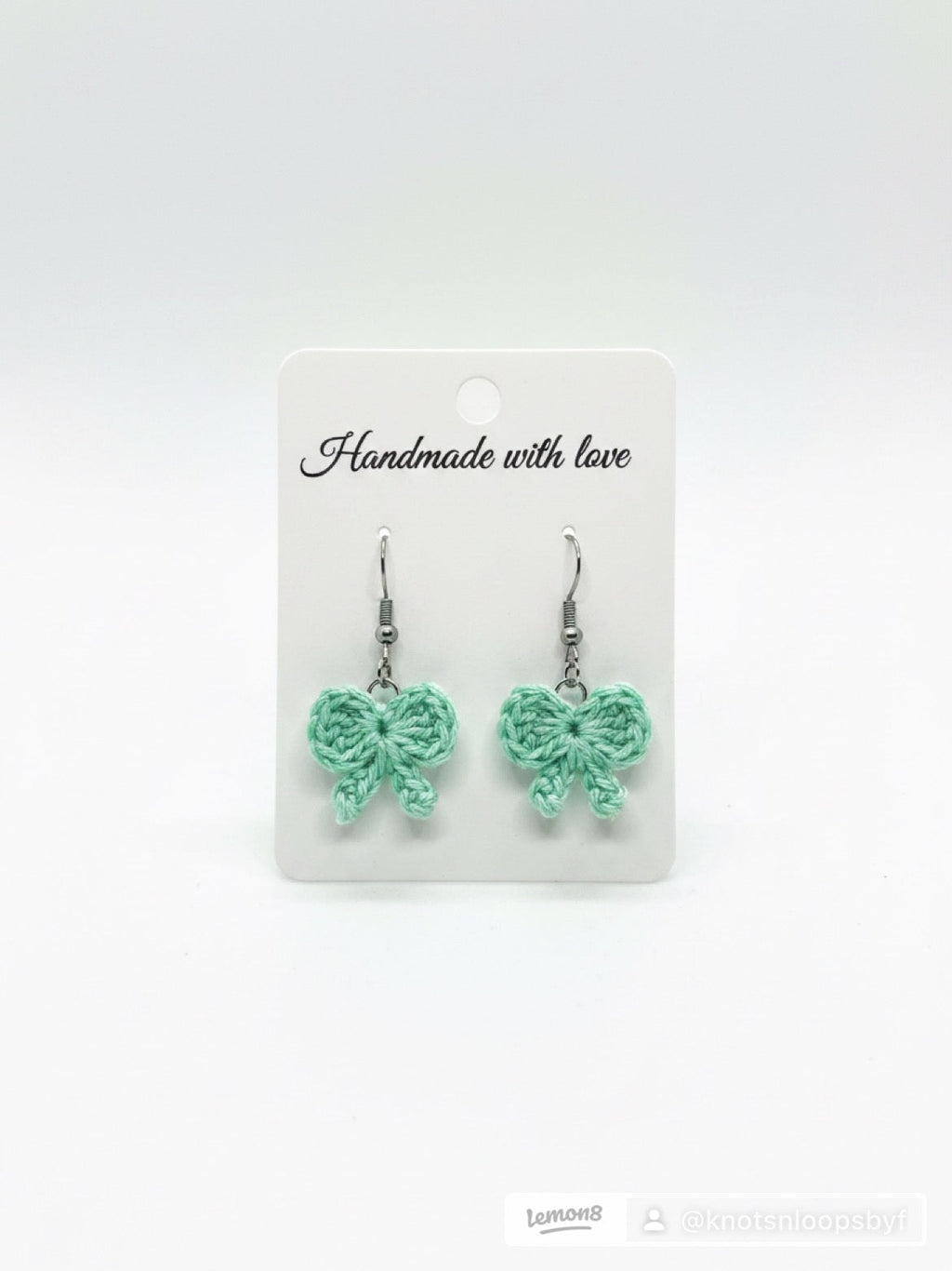 A pair of mint green crocheted bow drop earrings on a white display card that reads 'Handmade with love'.