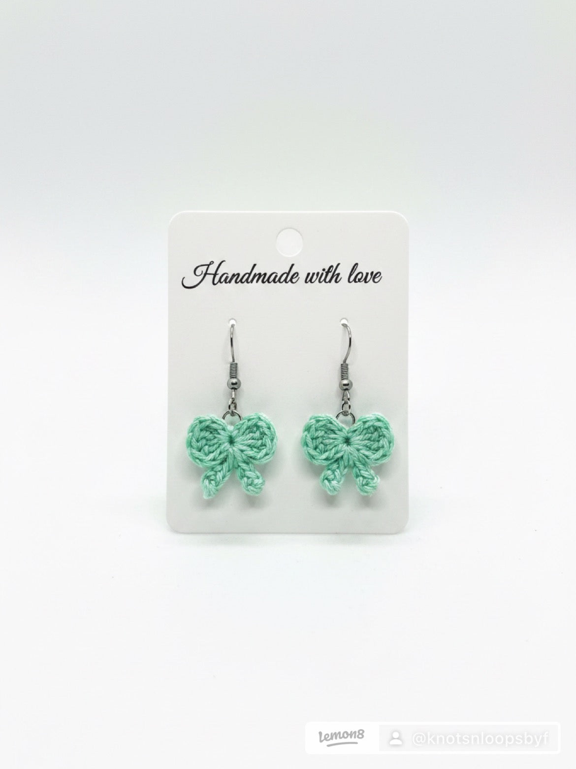 A pair of mint green crocheted bow drop earrings on a white display card that reads 'Handmade with love'.