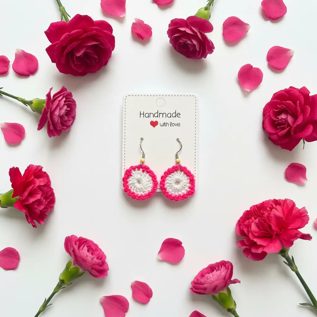 A pair of handmade crocheted hoop earrings with pink and white floral designs, displayed on a white card labeled 'Handmade with love' and surrounded by pink flowers and petals on a white background.