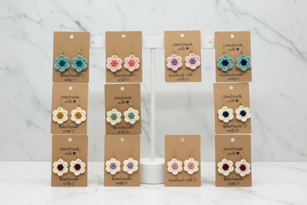 A set of handmade crocheted flower earrings displayed on brown kraft cards on a white stand against a light marble background. The earrings feature various colorful crochet flowers in blue, pink, yellow, green, purple, and red.