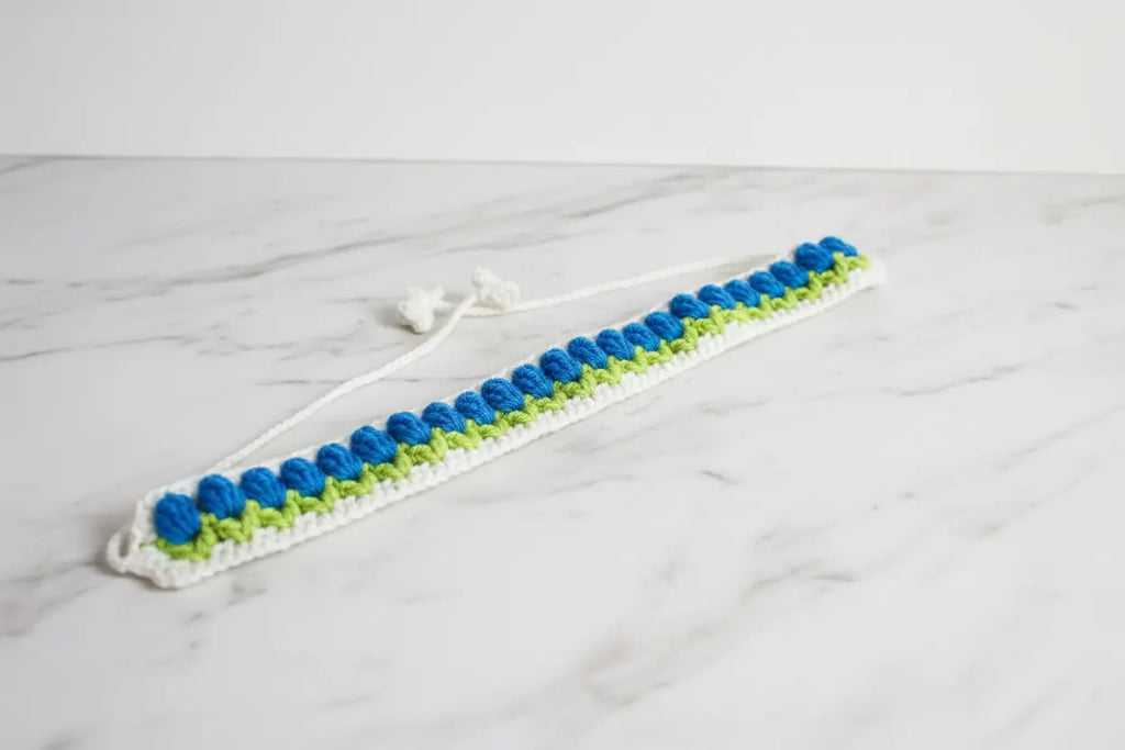 A handmade crochet hair claw clip made of white, blue, and lime green yarn, resting on a white marble surface.