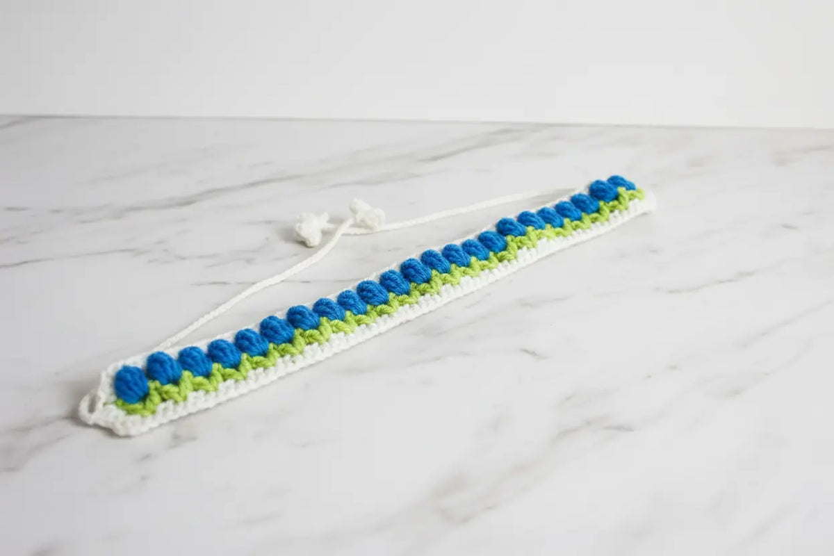 A handmade crochet hair claw clip made of white, blue, and lime green yarn, resting on a white marble surface.