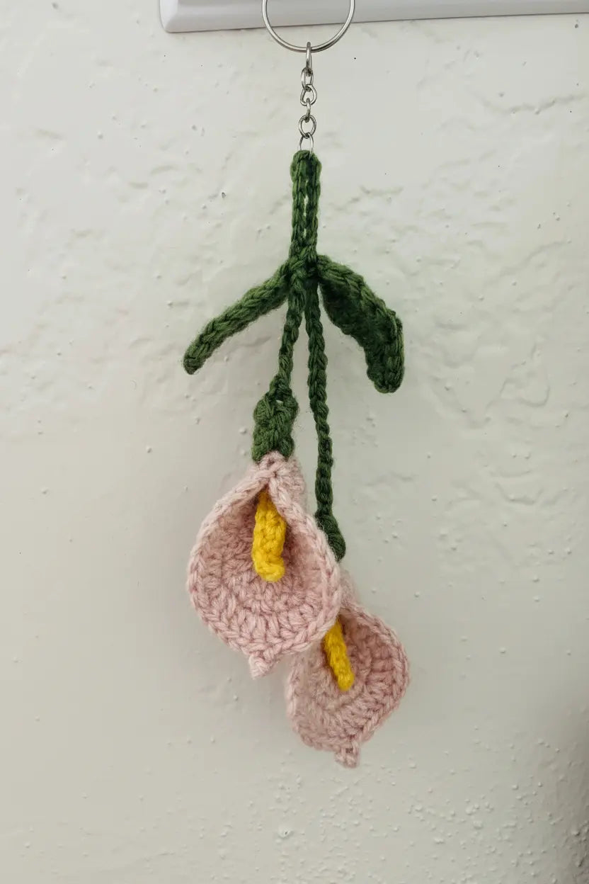 Handmade crocheted calla lilies with pink petals, yellow centers, and green stems hanging from a metal ring against a textured off-white wall.