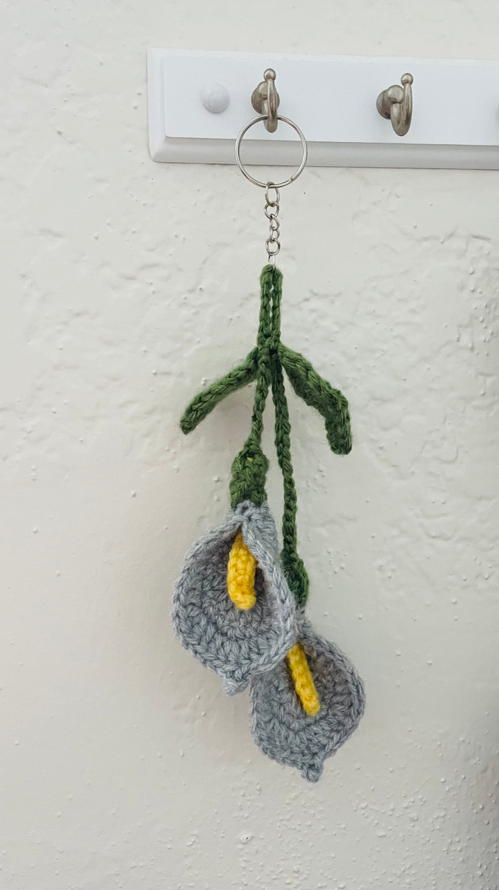 A handmade crocheted keychain featuring two gray calla lilies with yellow centers and green stems, hanging from a metal key ring on a white wall hook.
