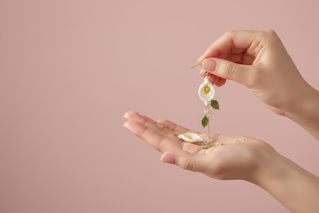 A close-up of a person's hands holding a delicate gold earring with a white daisy flower and green leaves, with the matching earring resting in the palm below, against a solid dusty pink background.