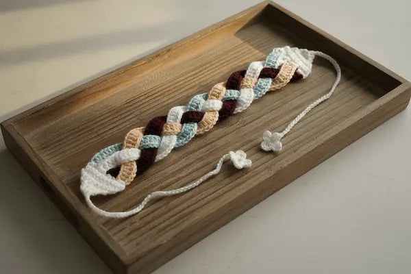 A crocheted braided bracelet with white, light blue, peach, and burgundy sections, displayed on a rectangular wooden tray.