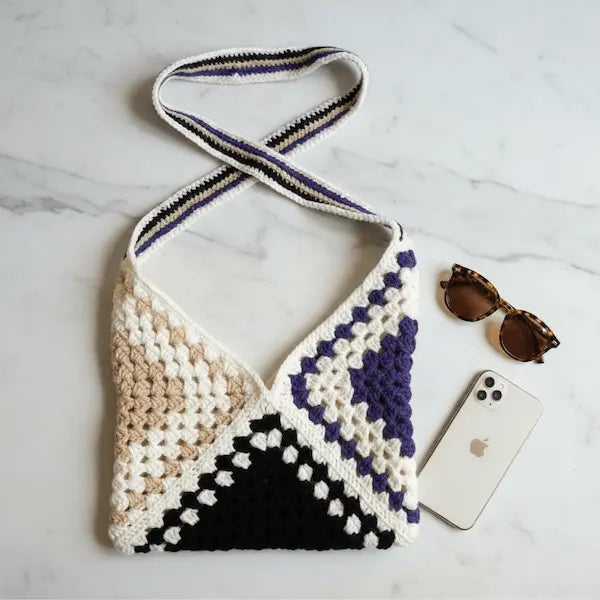 A handmade crocheted shoulder bag with a geometric triangle pattern in cream, beige, black, and purple, styled on a white marble surface with sunglasses, a smartphone, and a strap.