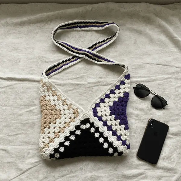 A handmade crocheted shoulder bag with a geometric pattern in black, white, beige, and purple, laid flat on a light beige fabric background next to sunglasses and a black smartphone.