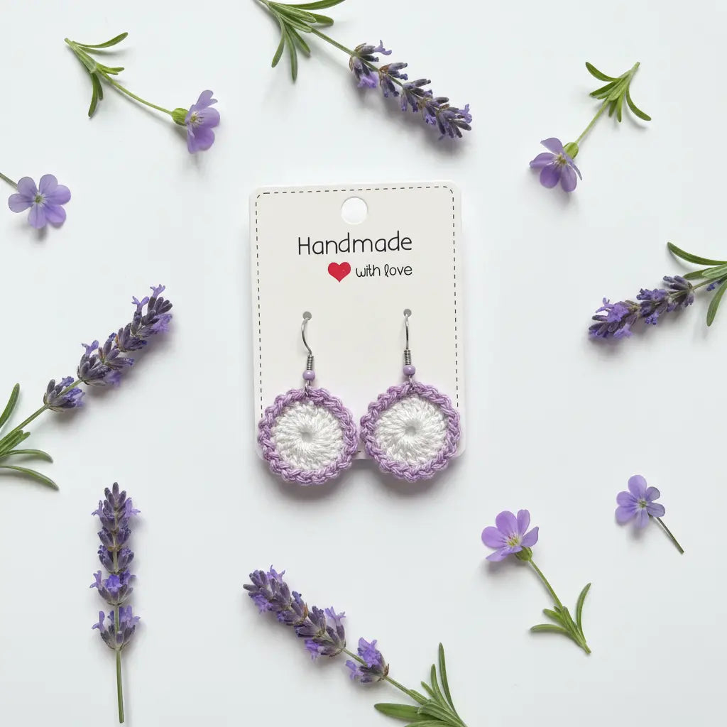A pair of handmade crocheted dangle earrings in white and lavender purple, displayed on a white backing card with 'Handmade with love' printed on it. The card is styled in a flat-lay arrangement with sprigs of lavender and rosemary on a clean white background.