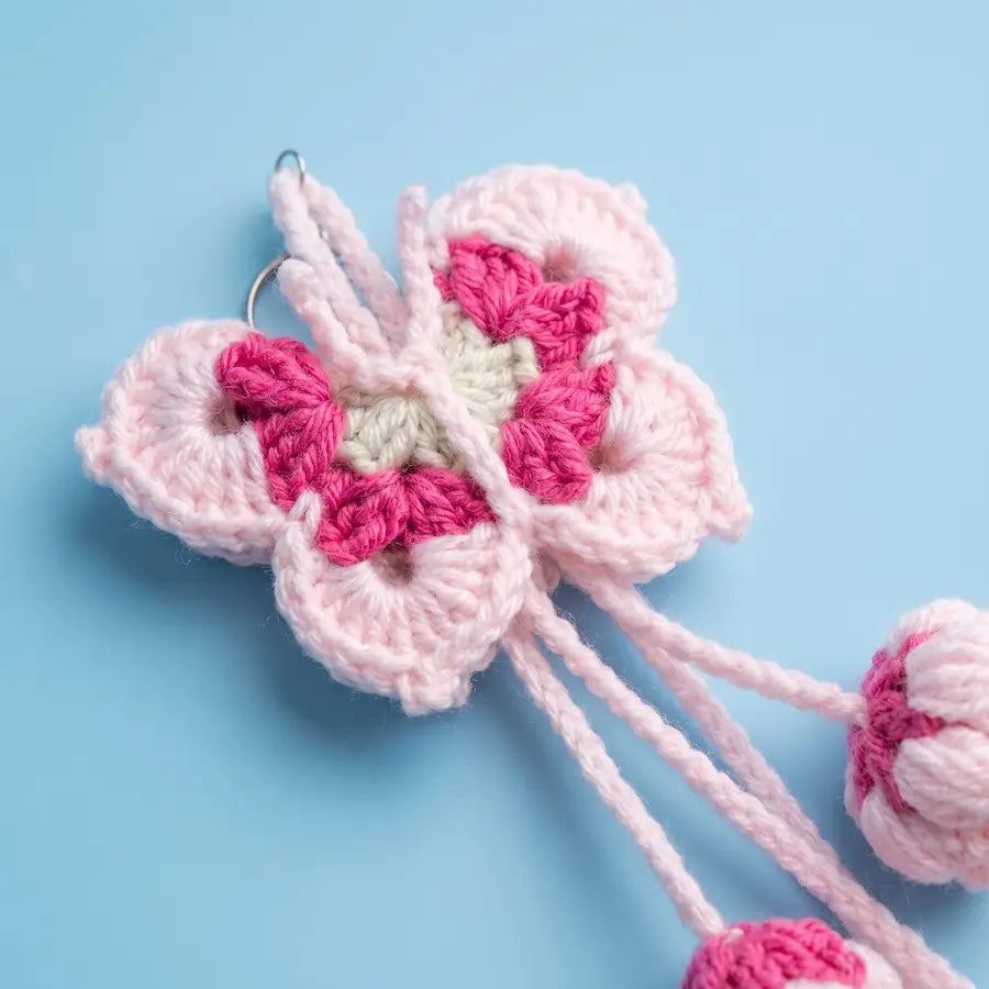 A crocheted flower-shaped hair clip in light pink, magenta pink, and cream, shown on a light blue background with its elastic bands visible.