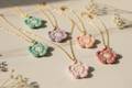 Five handmade crochet flower necklaces with gold chains are arranged diagonally on a light marble surface with dried flowers. The necklaces feature pastel green, lavender, peach, and dusty pink flowers with white centers.