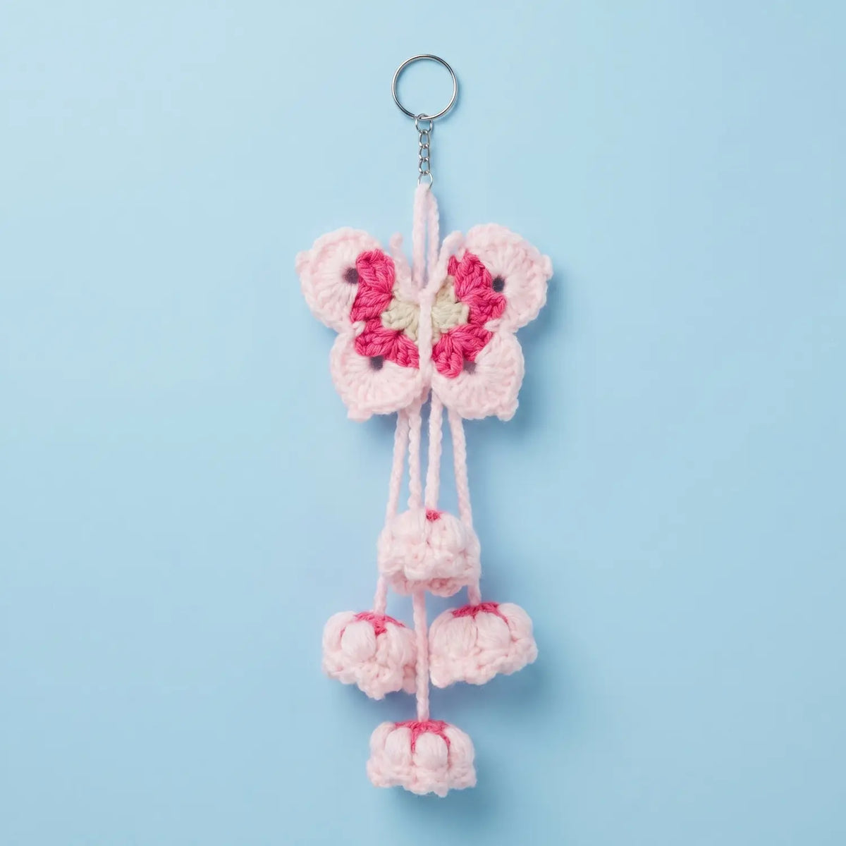 A crocheted cherry blossom keychain featuring a light pink butterfly with a bright pink and cream center, hanging from a silver metal key ring against a solid light blue background.