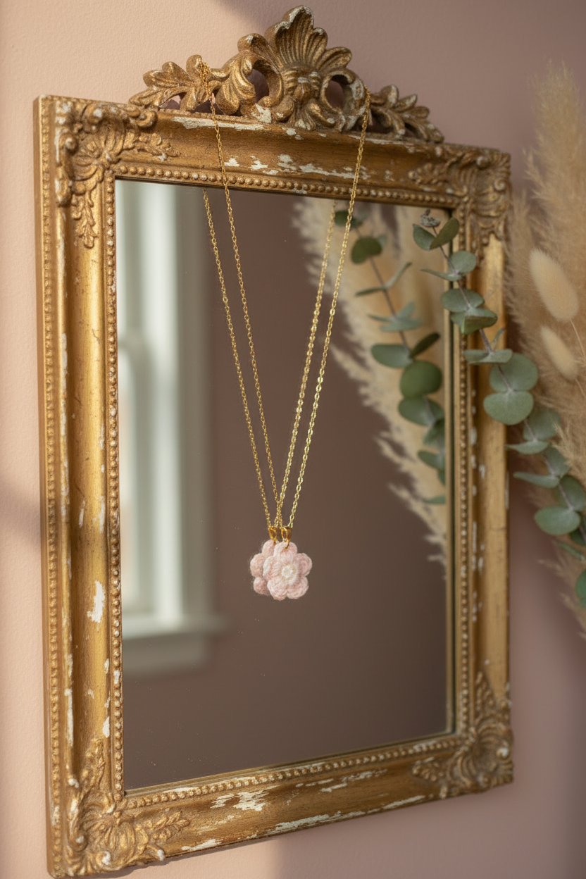 A gold ornate wall mirror with a pink flower pendant necklace hanging in front of it, set against a soft blush pink wall with dried grass and eucalyptus decor.