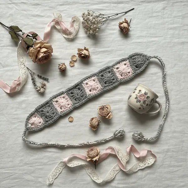 A crocheted eye mask with alternating gray and light pink granny squares, styled in a flat lay with dried roses, lace ribbons, and a small floral teacup on a white fabric background.