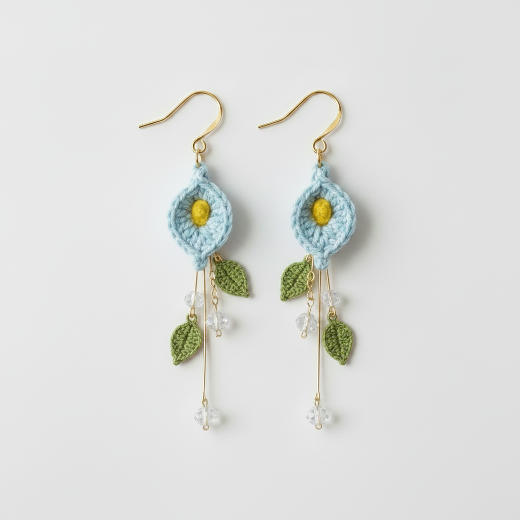 A pair of handmade crochet dangle earrings featuring light blue flower motifs with yellow centers, green leaves, and gold-tone hooks on a light gray background.