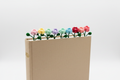 A beige hardcover notebook or journal standing upright on a white studio background, with a row of small crocheted flowers in pink, yellow, green, purple, blue, and red tucked along the top edge.