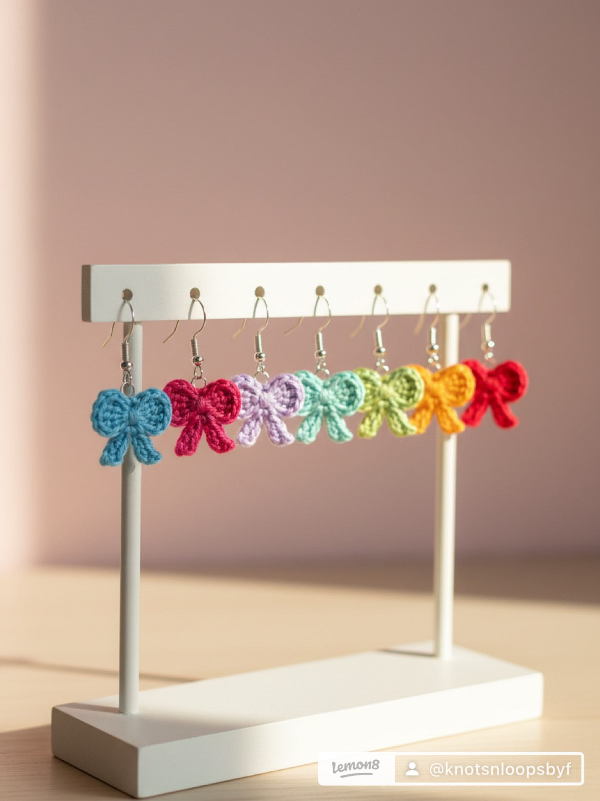 A set of colorful crocheted bow earrings displayed on a white stand against a soft pink background.