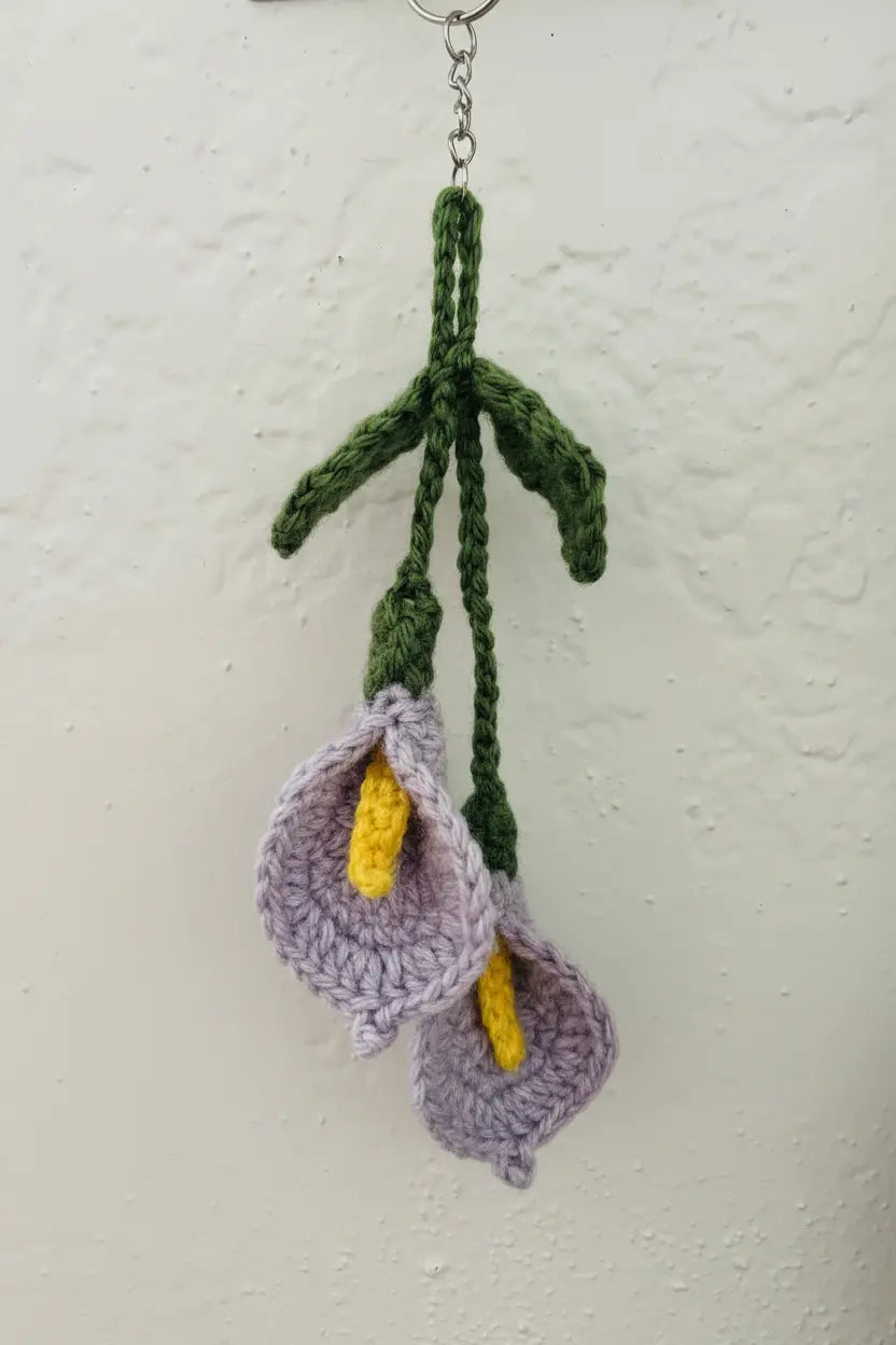 Handmade crocheted calla lily keychain hanging against a textured off-white wall. The keychain features two lavender crocheted calla flowers with bright yellow centers and dark green stems and leaves attached to a silver metal ring.