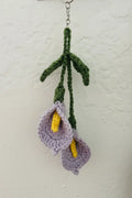 Handmade crocheted calla lily keychain hanging against a textured off-white wall. The keychain features two lavender crocheted calla flowers with bright yellow centers and dark green stems and leaves attached to a silver metal ring.