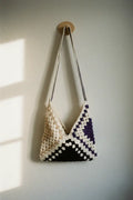 A handmade crochet shoulder bag with a geometric pattern in cream, beige, black, and purple, hanging from a wooden peg on a textured off-white wall.