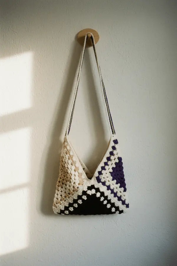 A handmade crochet shoulder bag with a geometric pattern in cream, beige, black, and purple, hanging from a wooden peg on a textured off-white wall.