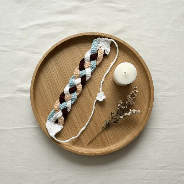 A crocheted braided bookmark with a white cord and small flower charm, displayed on a round wooden tray with a white candle and dried flowers on a light fabric background.