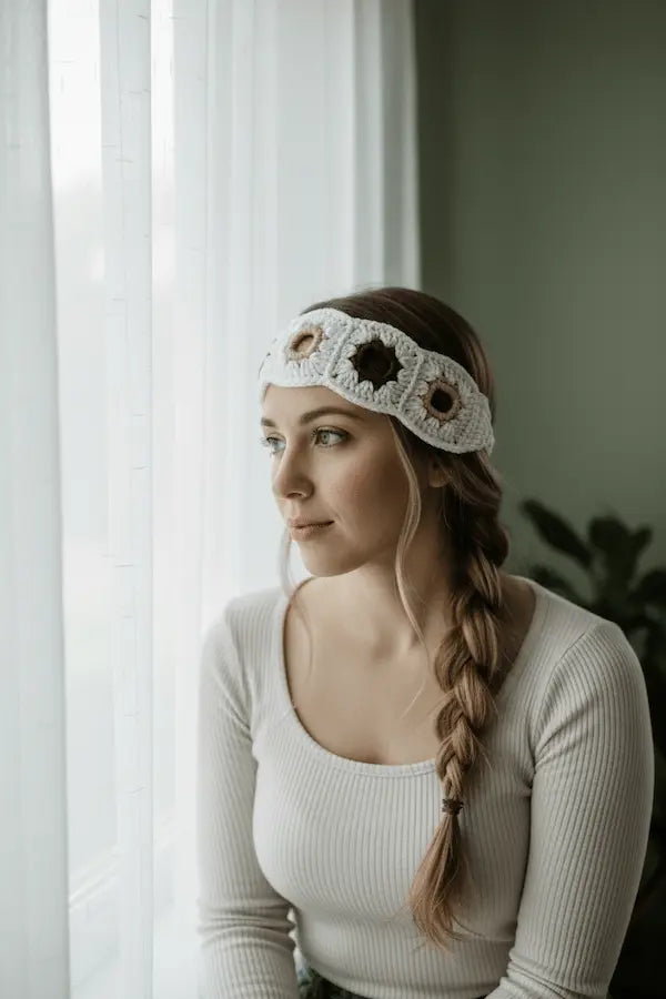 A woman with a long braid wearing a white crochet headband with brown star details, sitting by a window in a neutral-toned room.