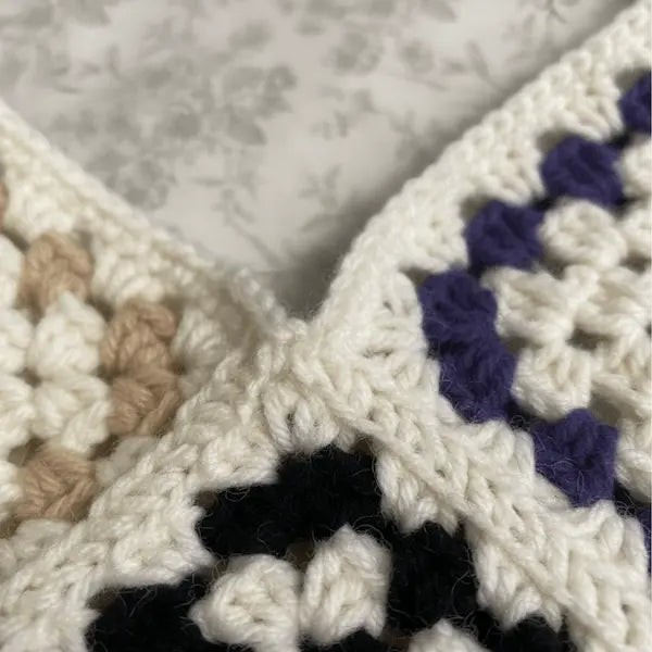 Close-up of a crocheted garment corner showing cream yarn, purple yarn, black yarn, and tan yarn in a granny square pattern against a light floral background.