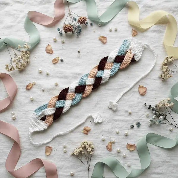 A handmade braided crochet bracelet with white, light blue, peach, and dark brown stripes, styled on a white textured fabric with pastel ribbons and dried flowers.