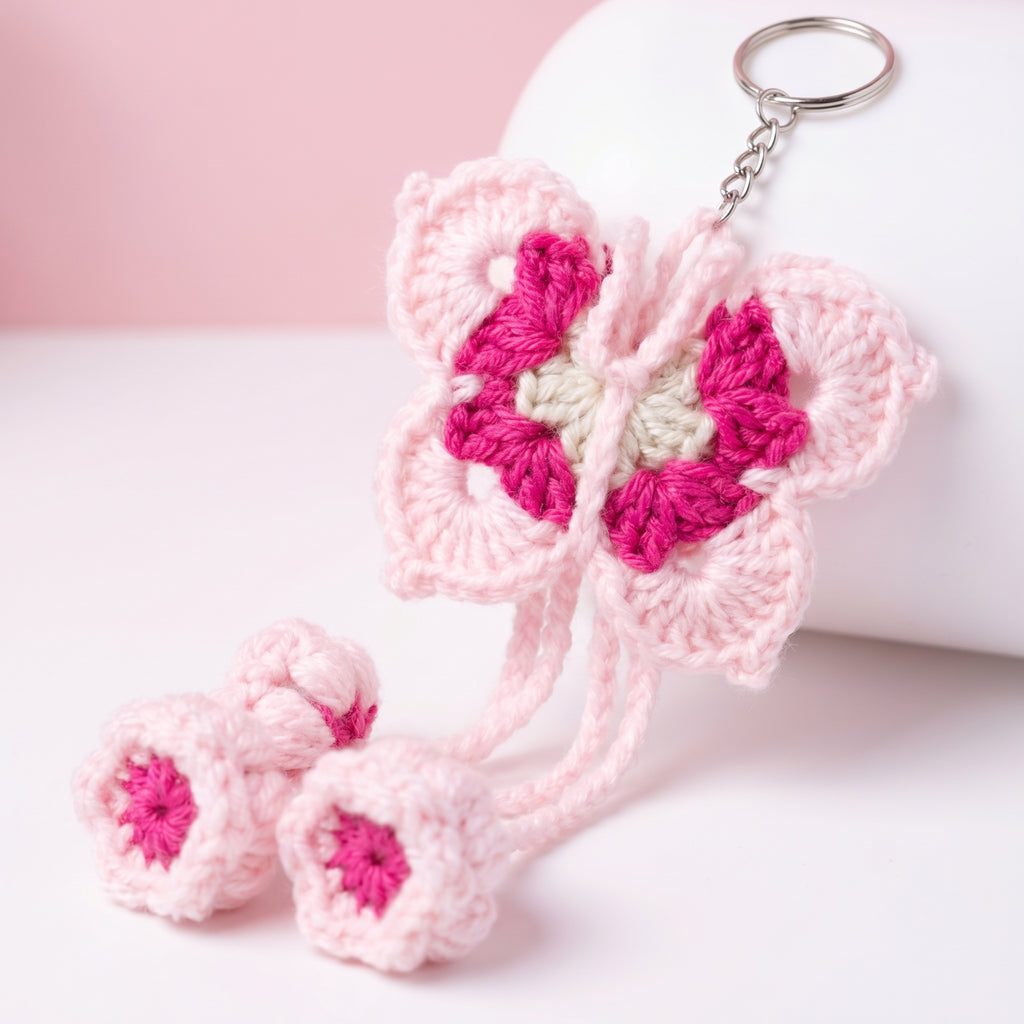A pink and magenta crocheted butterfly keychain with two matching flower charms, displayed on a white cylindrical stand against a soft pink and white background.